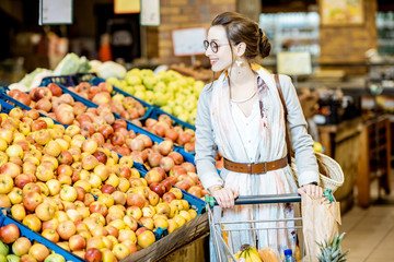 Young woman buying food, standing with shopping cart full of products in the supermarket with fruits on the background