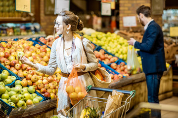 Woman packing apples into the eco bag with man holding plastic bag on the background in the supermarket