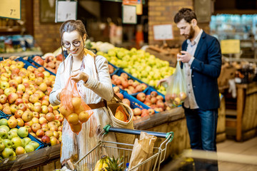 Woman packing apples into the eco bag with man holding plastic bag on the background in the supermarket