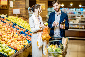 Young and happy couple buying fresh fruits and wine standing together with shopping cart in the supermarket