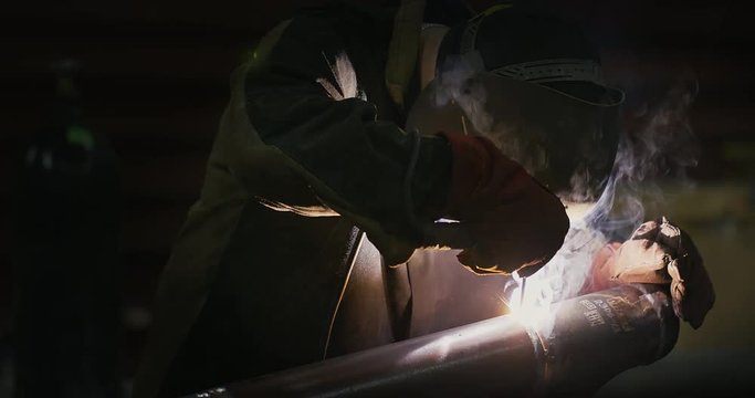 Welder in welding mask, in working clothes at the factory, welds the pipe in the factory room. 