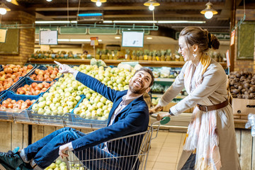 Man and woman having fun while riding with shopping cart in the supermarket with fruits and vegetables on the background