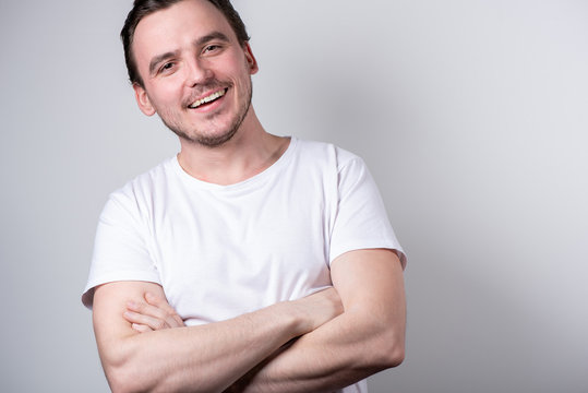Handsome brunette man in a white T-shirt with arms crossed is smiling on a gray background
