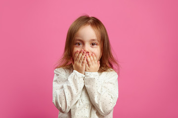 Little girl with a surprised look covered her mouth with hands and looks at you on pink isolated...