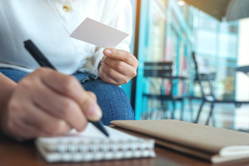 Closeup image of a woman holding a blank empty business card while writing on a notebook