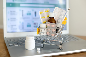 Shopping cart toy with medicaments in front of laptop screen with pharmacy web site on it. Pills, blister packs, medical bottles, thermometer set. Health care and internet shopping. - 255332146