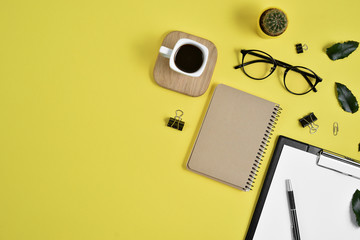 Office desk workspace with blank clip board, office supplies, pen, cactus, green leaf, coffee cup on a wooden stand and eye glasses on yellow background. Flat lay, top view blog concept