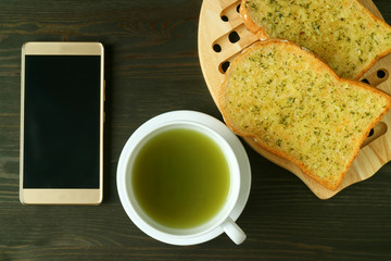 Empty Screen Smartphone with a cup of Hot Green Tea and Garlic Butter Toasts on Dark Color Background
