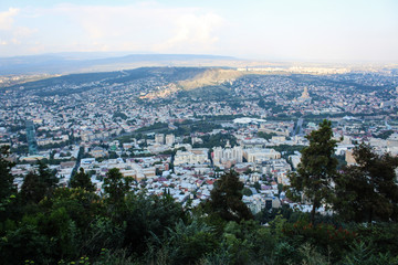Obraz premium The capital of Georgia is Tbilisi, from a height. View from the observation deck in the park Mtatsminda. Big city. Houses, buildings, churches, the Kura River. Greens, trees, shrubs, summer.