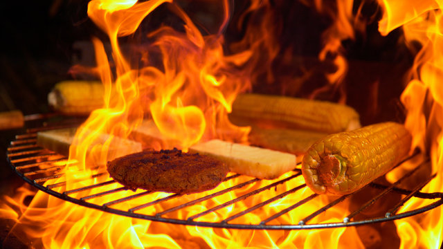 MACRO: Flames engulf vegetarian patties, corn, and cheese grilling during picnic