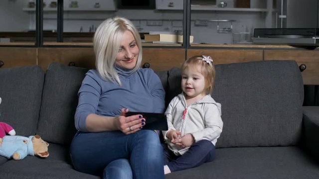 Positive mother and cute little daughter watching developing video on smart phone sitting on sofa in living room. Joyful child looking at phone screen while making online easy educational exercises.