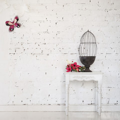 Obraz premium Vintage wooden table with metal cage against the background of a white brick wall. Paper flowers on the table and a butterfly on the wall. Minimalistic conceptual photozone.