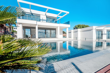 Fototapeta na wymiar Luxury modern white house with large windows overlooking a Mediterian landscaped garden with palm trees and blue swimming pool. High tech style villa. Vacation home or hotel. Modern loft design.ees a