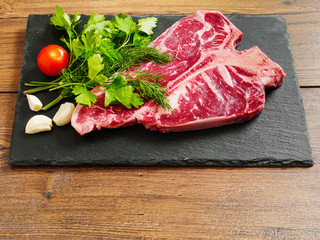 T bone steak uncooked on a stone plate with herbs, garlic cloves and tomato.