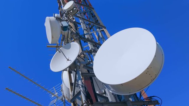 High telecommunication tower standing on top of a mountain against the blue sky with clouds. On the mast there are satellite dishes, sector panel antennas, a radio relay dishes, blocks of the radio 
