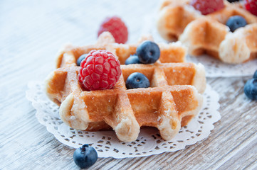 Traditional waffles with fresh  raspberries and blueberries on lace doily on wooden background. - obrazy, fototapety, plakaty