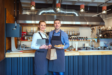Smiling young man and woman using tablet at small eatery restaurant – Happy business owners in apron holding paper bag with to go order from online customers - 255344769