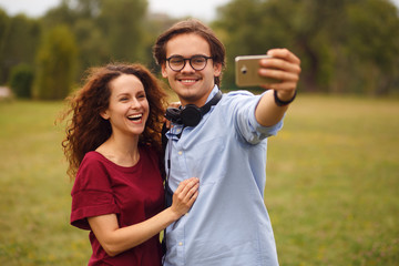 Two young smiling person, doing selfie in public park, isolated on a green grass in open air. Leisure time. Horizontal view.
