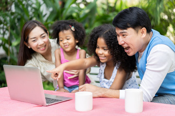 Happy family checking their social media or video call on a laptop computer as they relax together on backyard. parents or Volunteers take care of orphans pretty little daughter in child care center