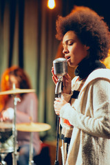 Mixed race woman singing. In background band playing instruments. Home studio interior.