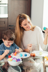 Blonde Female babysitter having confidence, skill and understanding to support children's play, interacting with boy in a playful way, painting eggs to Easter celebration.