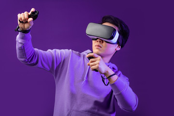 Joyful Japanese youngster playing videogame in virtual reality helmet on dark blue background. Smartphone using with virtual reality goggles. Technology, simulation, hi-tech, game concept