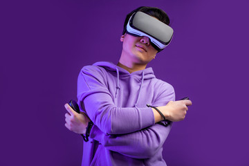 Young Asian guy looking proud body during watching sport game using virtual reality headset, posing isolated over dark violet background.