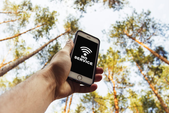 Male hand is holding a phone with sign no service  above his head against the background of trees in a pine forest. Concept of no service, wifi, internet in the forest or a place far from the city