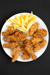 Tasty fried chicken drumsticks, spicy wings, French fries, chicken tenders on white plate over black background, top view. Flat lay, overhead, from above.
