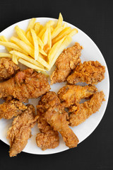 Tasty fastfood: fried chicken drumsticks, spicy wings, French fries, chicken strips on white plate over black surface, top view. Flat lay, overhead, from above.