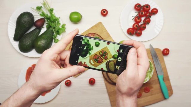 A man photographs on a smartphone sandwiches with avocados, cherry tomatoes and salmon fillets.