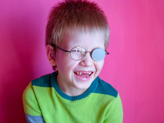 Portrait of funny child in new glasses with patch for correcting squint on pink background. boy with lost baby teeth..Ortopad Boys Eye Patches nozzle for glasses for treatment of strabismus (lazy eye)