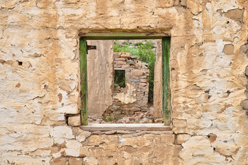 Fototapeta na wymiar Central empty green window frame on rocky wall of abandoned house. Through frame see crumbled walls of remaining ruined house