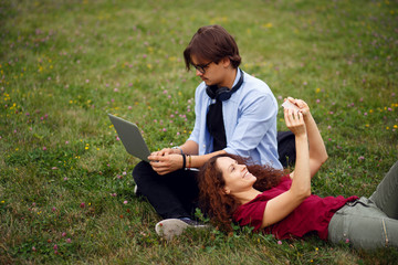 Two friends seated on a grass in urban park, using a laptop and smart phone for self-portrait, isolated green grass background, beautiful day for works quietly in open air.