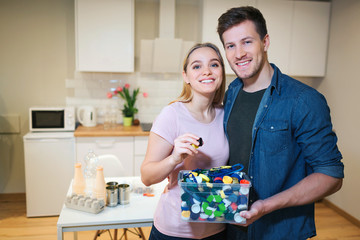 Recycling. Young smiling couple holding plastic lids in the container with green recycle icon on kitchen background