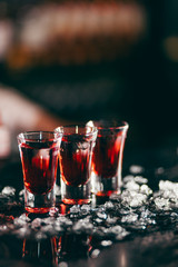 Three red alcohol shots standing in line surrounded with ice pieces, isolated on dark blurred background. Concept of alcohol, spirits for a good night time.