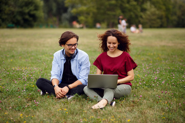 Two happy friends seated on a grass in urban park, using a laptop, isolated on a green grass forest background, beautiful day for works in open air. Communication, relationship,friendly concept.