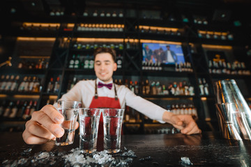Close-up of expert bartender working in a bar. Focus on shots with vodka or another strong alcohol, served by smiling pleasant barkeeper.