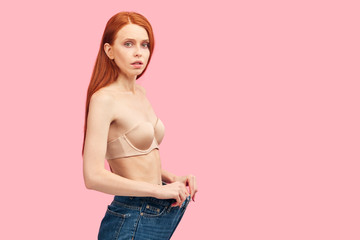 Young exhausted depressed sad red-haired woman having some problems with eating habbits pulls oversized pants down jeans showing her diet results against pink wall
