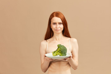 Symptoms of anorexia manifested in disgust for food. Portrait of grimacing unsatisfied facial emotional expression young woman refusing to eat fresh broccoli on plate, posing over pink background