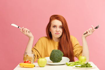 Puzzled, uncertain young red-haired woman sitting at table, served with broccoli shrugging with...