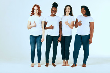 Obraz premium Four young multi ethnic female models of different race, body size and hair color smiling at camera, show Thumb up on white studio background in white casual t-shirts and skinny jeans.