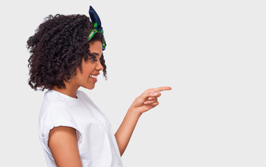 Side view portrait of happy African American young woman dressed in white t shirt and green headband, indicates aside with forefinger to blank copy space, isolated over white wall.