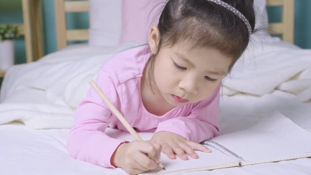 Happy child little asian girl writing in a book on the bed in the bedroom, Educational concept for homeschool