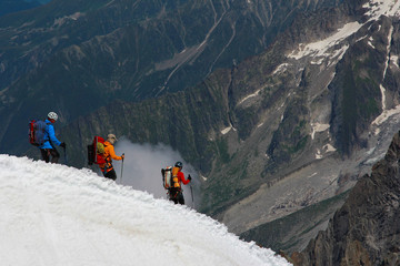 A group of three-person climbers continuing their descent along a ridge against the backdrop of mountains and clouds. Team work concept