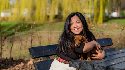 Loving scene of a mature woman hugging her dachshund puppy who is sitting on a wooden picnic table, blurred green background, long black straight hair, casual clothes, sunny day to enjoy in the park