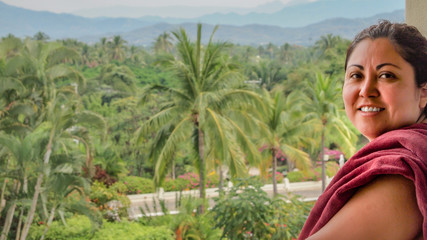 Partial portrait of smiling middle-aged Mexican woman standing on balcony against green palm trees in blurred background, looking at camera, hair tied back, enjoying vacation in tropical day weather