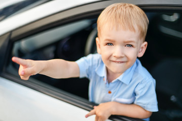 A small blond-haired blue-eyed boy in the front seat of the car shows the index finger of his right hand. Happy kid behind the wheel of a big car