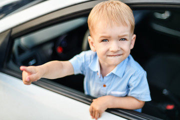A small blond-haired blue-eyed boy in the front seat of the car shows the index finger of his right hand. Happy kid behind the wheel of a big car