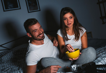 Young couple having fun watching television at night dressed in pajamas and eating popcorn.